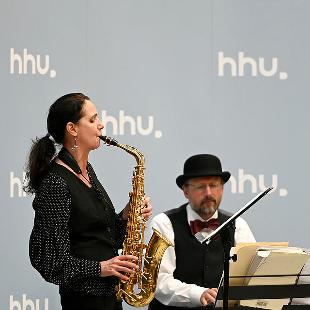 Zwei Musiker vor einer HHU-Fotowand. Links eine Frau mit Saxophon, rechts ein Mann am E-Piano.