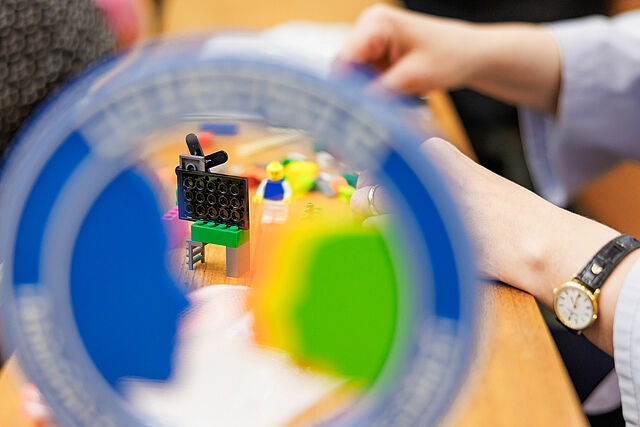  Eine Lehrpreis-Trophäe steht im Vordergrund. Im Hintergrund des Fotos baut eine Nominierte mit Lego Serious Play eine Szene aus ihrer Lehre nach.